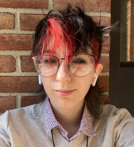 a person against a brick wall. the are white, with black and red hair, wearing a sweater with a collared shirt underneath. they have eyeliner and red eyeshadow on, as well as airpods in their ears.